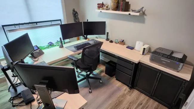 Modern home office with multiple monitors, ergonomic chair, printer, and decorative shelves on wood floor.