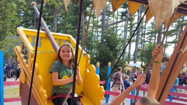 Two children smiling and swinging on a pirate ship ride at an outdoor fair with trees in the background
