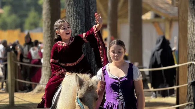 Girl in medieval dress leads a pony with a child rider waving at a Renaissance fair among tall pine trees.