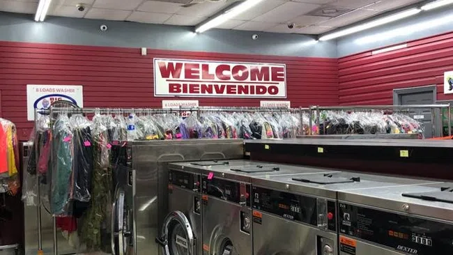 Interior of a laundromat with stainless steel washers and racks of clothes under a Welcome Bienvenido sign.
