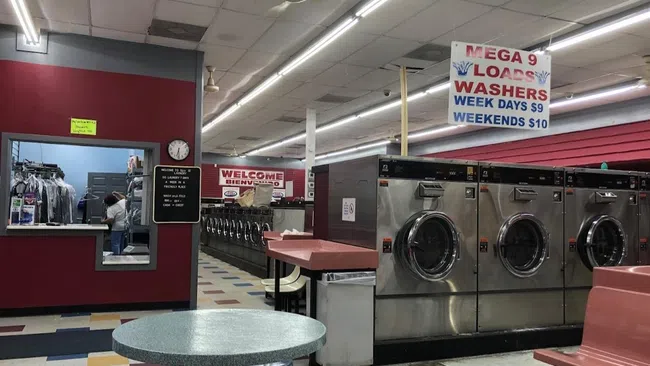 Interior of a laundromat with washing machines, colorful tiled floor, seating, and pricing signs for loads and washers.