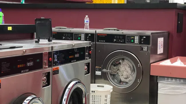 Interior of a laundromat with stainless steel washers, a laundry basket, trash bin, and pricing sign in the background.