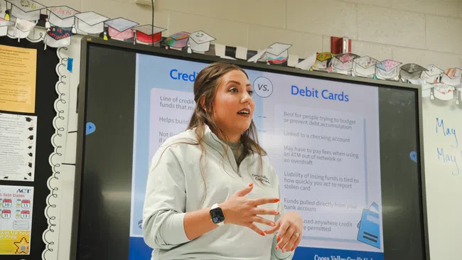Teacher explaining credit cards versus debit cards to students with an educational display in the background.