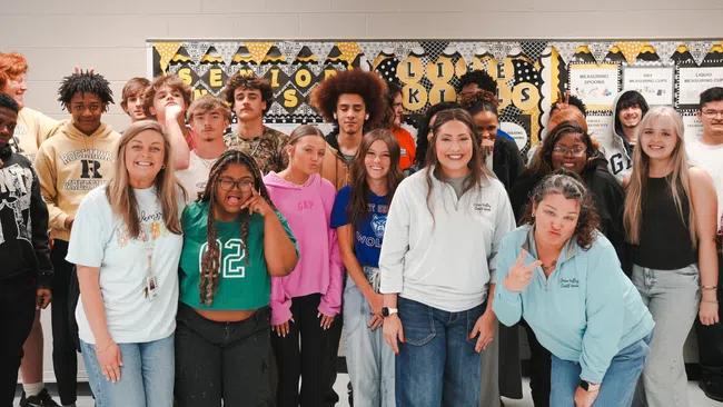 Group of diverse high school students and teachers smiling together in a classroom with decorated walls.