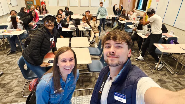 Diverse high school students and teacher smiling and posing for a selfie in a bright classroom setting.
