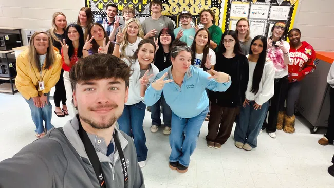 Group of smiling high school seniors posing for a selfie in classroom with Senior Life Skills bulletin board in background