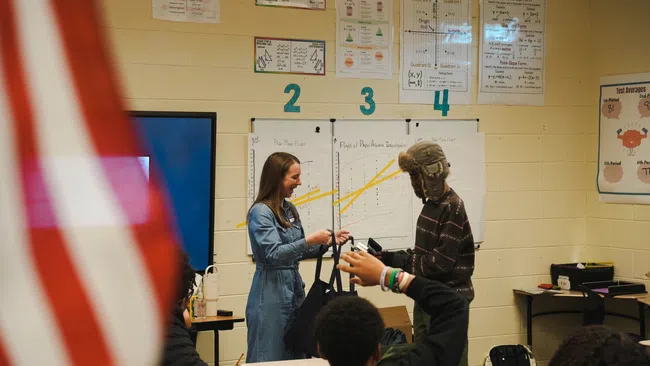 Teacher interacting with a student in a classroom with math posters and an American flag partially visible