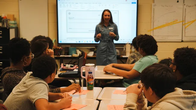 Teacher presenting to a group of attentive students in a classroom with charts and a digital display