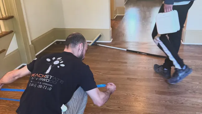Worker kneeling applies blue painter's tape on hardwood floor while another person walks holding a toolbox.