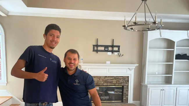 Two workers posing with knee pads in a room with wooden floor sample strips, fireplace, and chandelier.