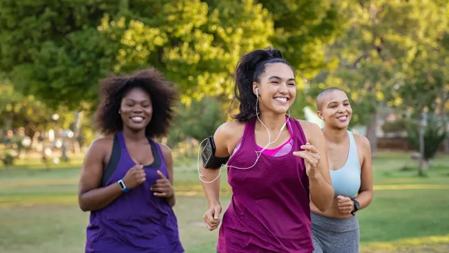 Three diverse women jogging outdoors in athletic wear, smiling and enjoying exercise in a green park.