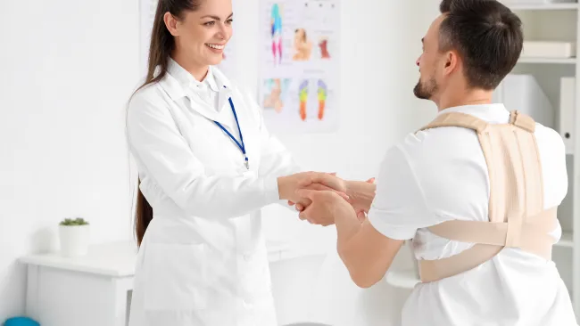 Smiling female doctor shakes hands with male patient wearing posture corrector in medical office.