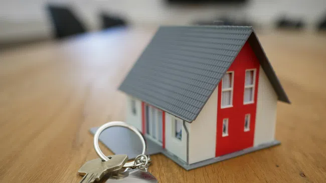 House key with a house-shaped keychain next to a small model house on a wooden surface.