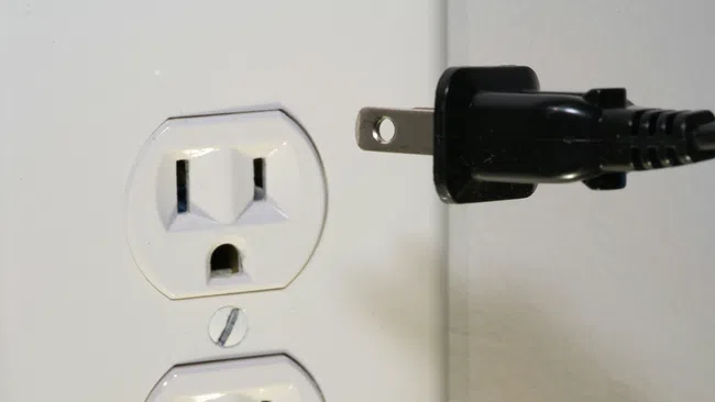 Black power plug about to be inserted into a white electrical outlet on a wall.