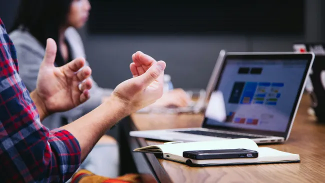 Person gesturing with hands during a meeting with a laptop, notebook, and smartphone on the wooden table.