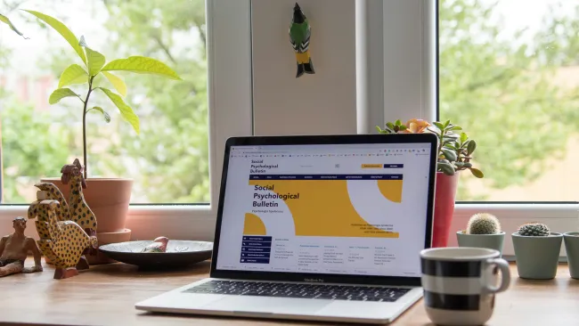 Laptop on wooden desk displaying Social Psychological Bulletin website near potted plants and striped coffee mug