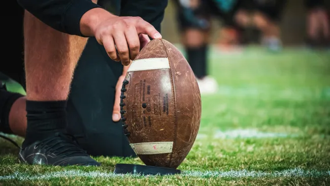 Close-up of a football player placing a worn football on a tee before a kick on a green field.