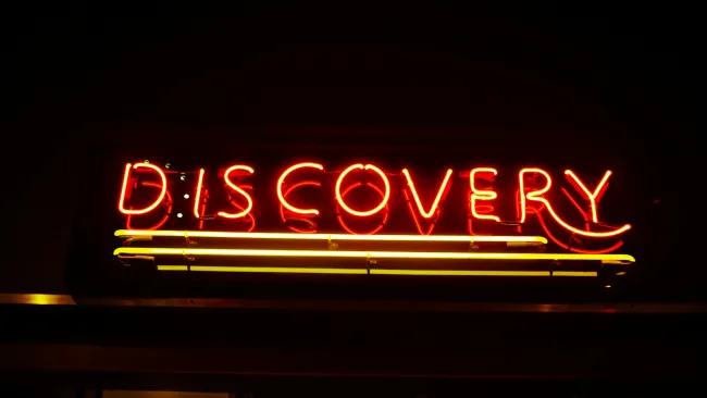 Red and yellow neon sign spelling DISCOVERY with two yellow horizontal lines below on a black background.