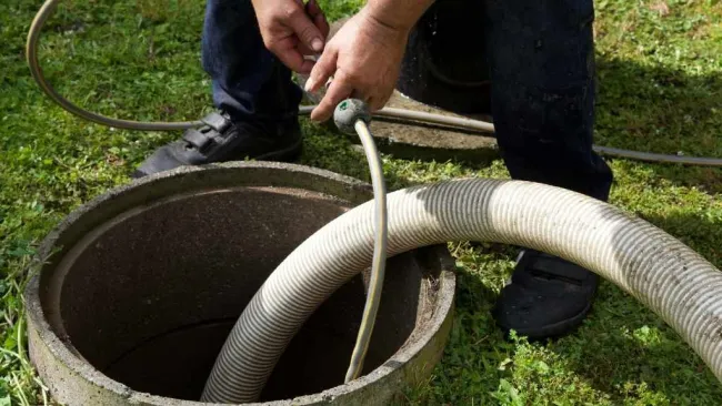 Worker handles hose inserted into a concrete septic tank opening on grassy ground outdoors.