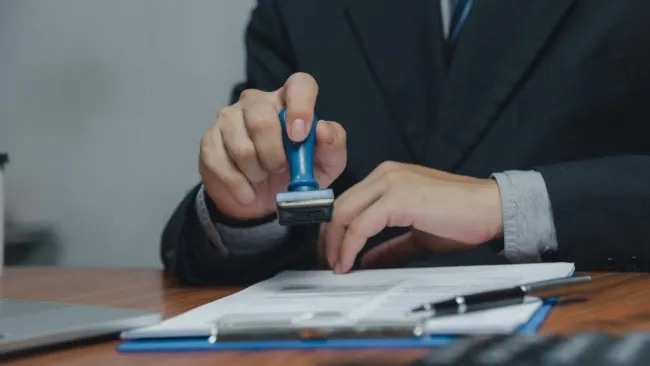 Businessman stamping a document on a desk with a laptop and pen nearby in an office setting.