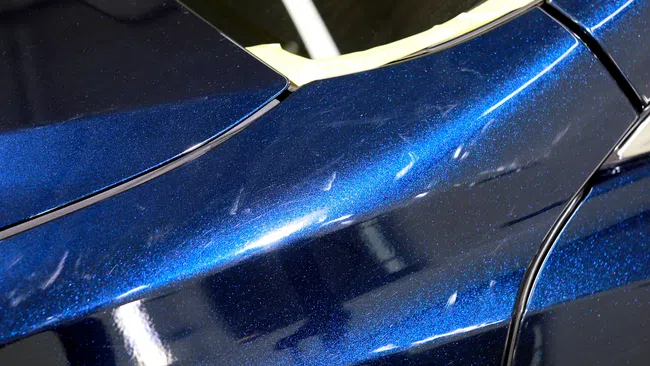 Close-up of blue metallic car paint with reflection and visible swirl marks on the surface.