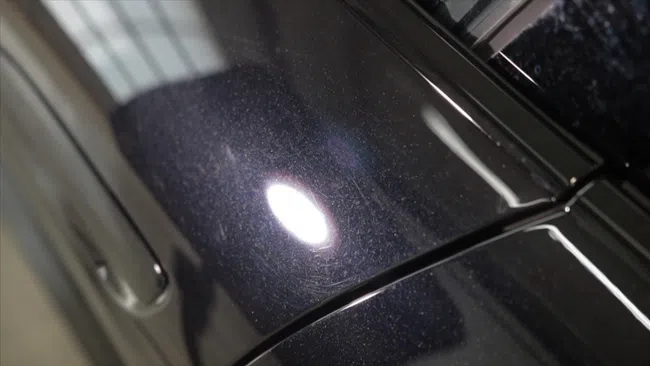 Close-up of black car surface with visible swirling scratches and light reflection showing paint imperfections.
