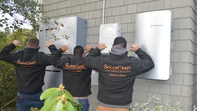 Three technicians wearing SolarCraft jackets stand in front of Enphase energy equipment installed on a house exterior.