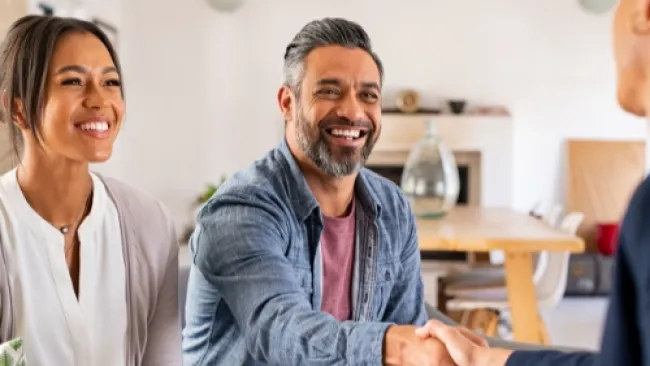 Smiling couple shaking hands with a professional in a cozy living room setting, holding documents and discussing plans.