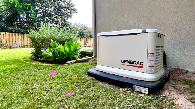 Generac standby generator installed outside a house on a grassy patch near garden plants and a wooden fence.