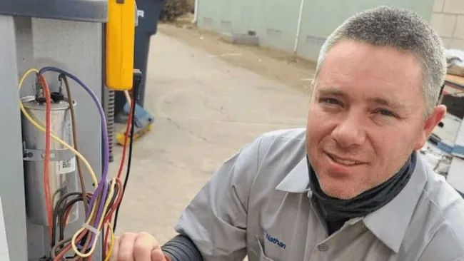 Technician testing electrical wiring on HVAC unit outdoors wearing a uniform and smartwatch.