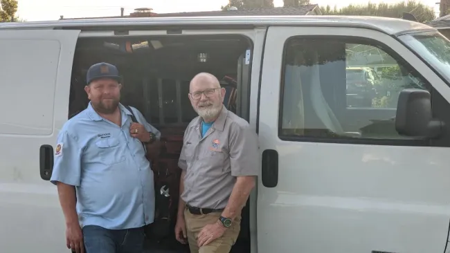 Two maintenance workers standing by a white work van with equipment inside on a sunny day