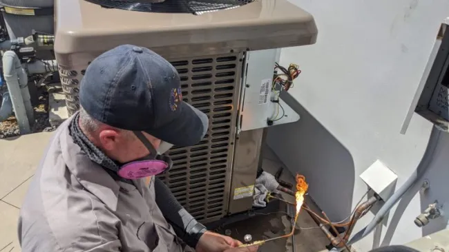 Technician wearing protective mask and cap soldering copper pipes next to an air conditioning unit outdoors.