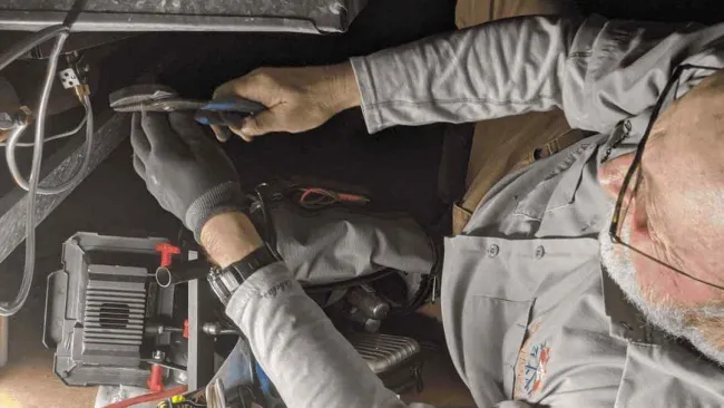 Electrician repairing wiring in a confined space with tools spread out on the floor under dim light.