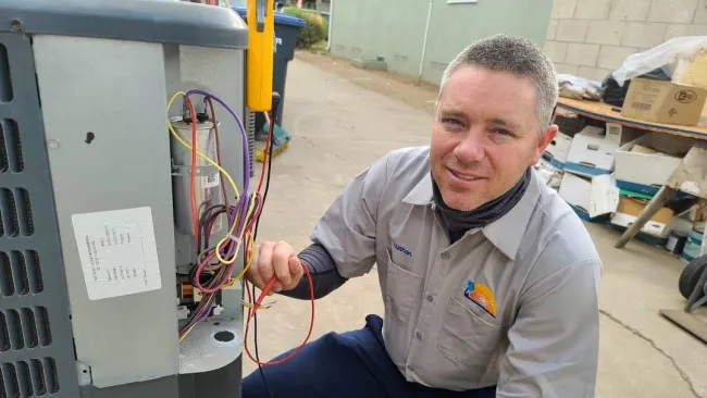 Technician in uniform troubleshooting HVAC unit wiring outdoors on a residential property.
