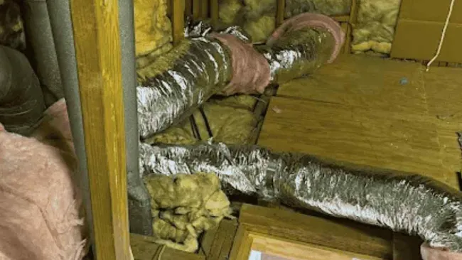 Attic space with exposed ductwork insulated with pink and yellow fiberglass and wooden framing around an attic access door.