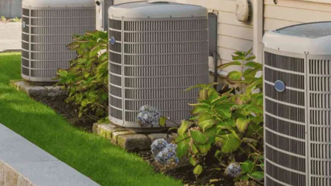 Three outdoor air conditioning units installed beside a house with plants and green grass nearby.