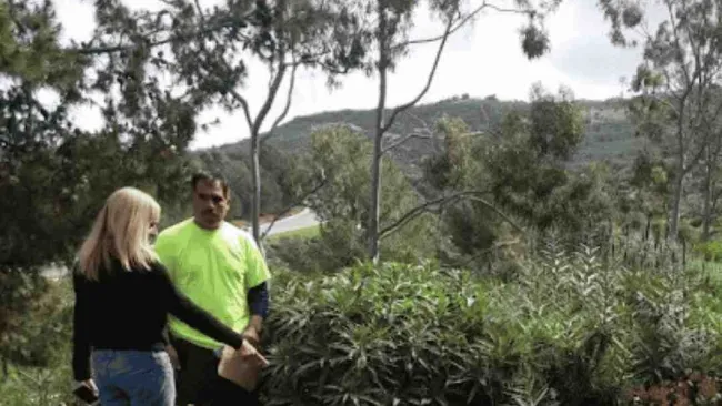 Two people discussing plants in a lush garden with shrubs and trees under an overcast sky.
