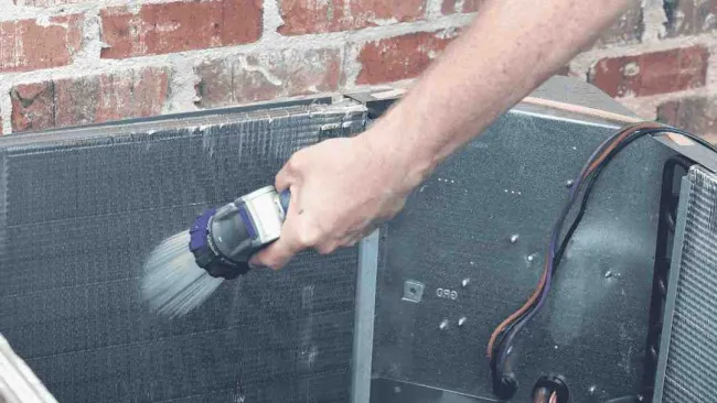 Technician cleaning an air conditioning unit's coil with a brush near a brick wall outdoors.
