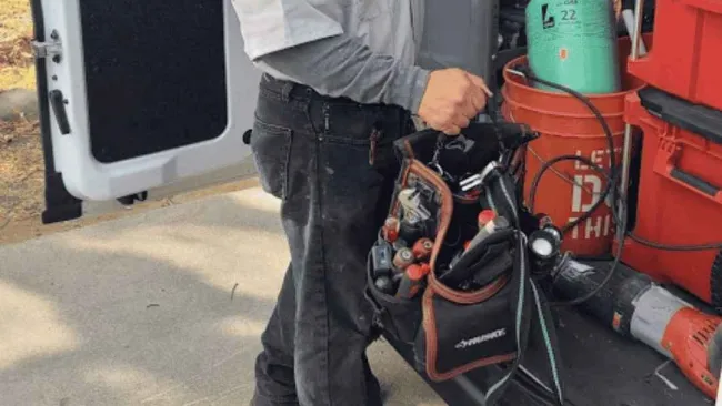 Technician wearing a face mask holding a tool bag next to a van loaded with equipment and supplies.