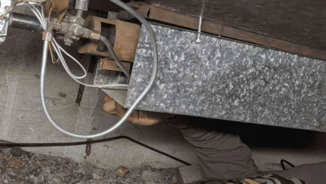 Technician inspecting and repairing HVAC ductwork in a confined basement space with tools and cables visible.