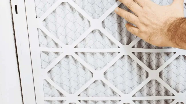 Hand replacing a white pleated air filter with a diamond pattern frame in an HVAC unit.