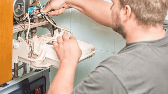 Technician repairing a wall-mounted heating boiler with tools in a domestic setting.