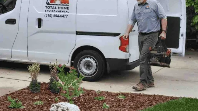 Man carrying toolbox walking from Total Home Environmental service van near landscaped yard with mulch and plants.