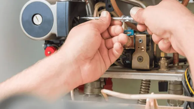 Hands using screwdriver to repair or maintain complex mechanical or heating system components indoors.