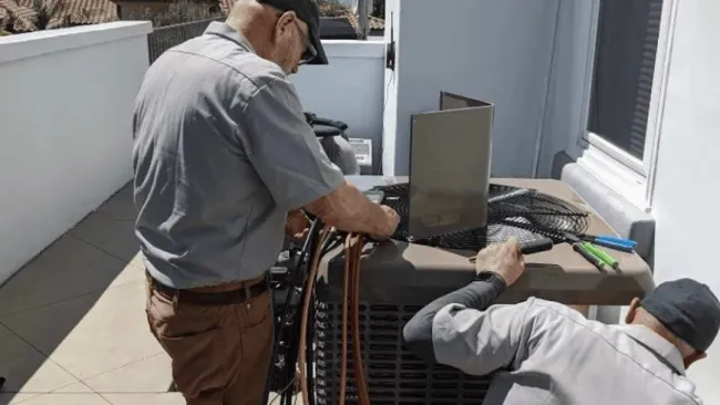 Two technicians servicing an outdoor air conditioning unit on a residential balcony under clear sky.