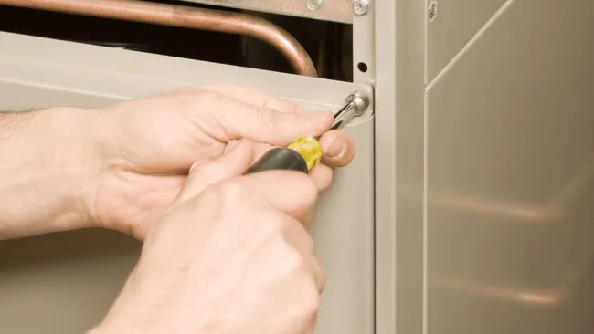 Hands using a screwdriver to tighten a screw on a metal HVAC unit panel during maintenance.