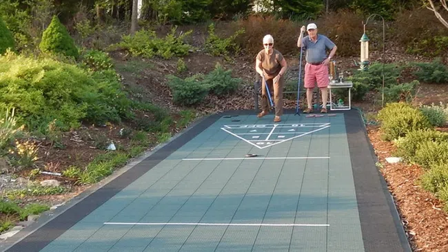 Two seniors playing shuffleboard on an outdoor court surrounded by lush greenery and bushes.