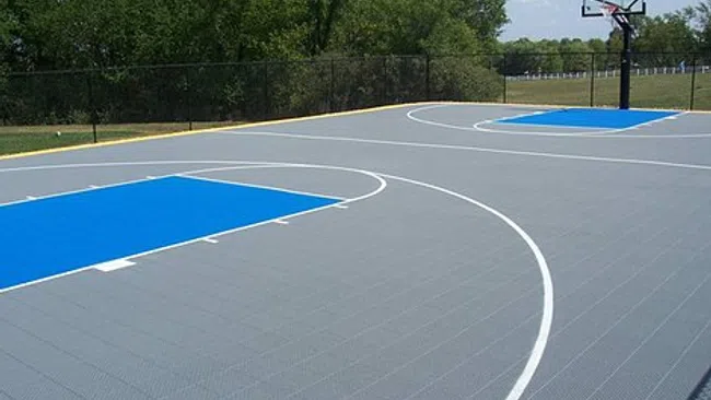 Outdoor basketball court with gray surface and blue key areas under clear sky and trees.