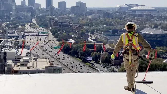 Construction worker in safety gear overlooking city skyline and busy highway from a high rooftop on a sunny day