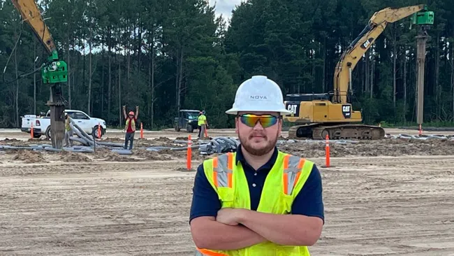 Construction worker in safety vest and helmet stands on site with excavators and forest background under blue sky.